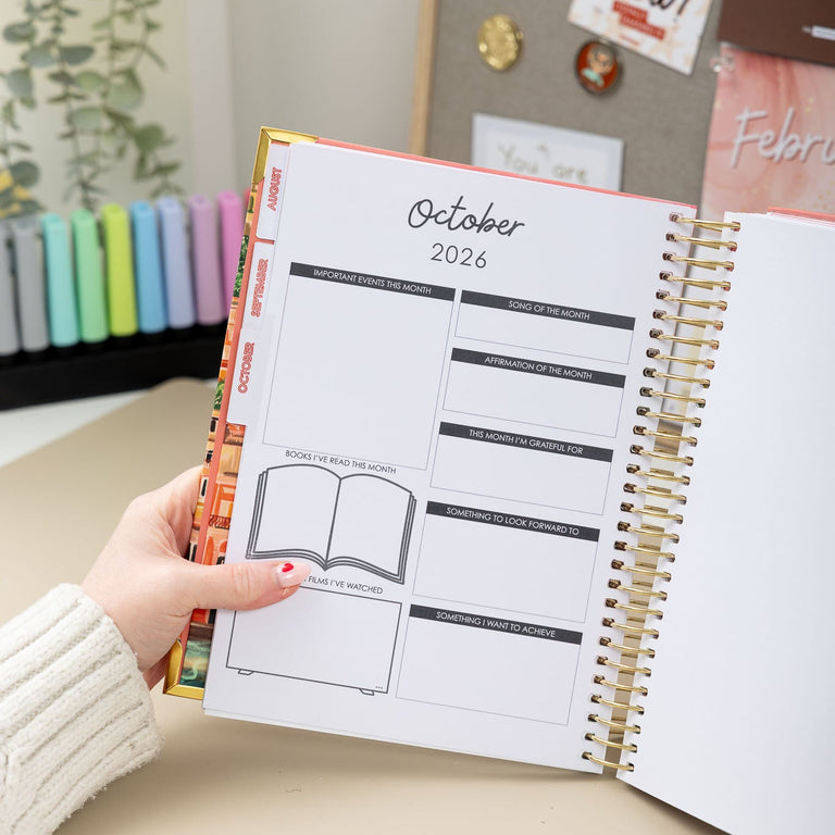 Person holding a planner open to October 2026 on a desk with decorative elements.