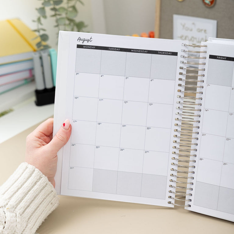 Person holding a spiral-bound planner with a calendar page on a desk.