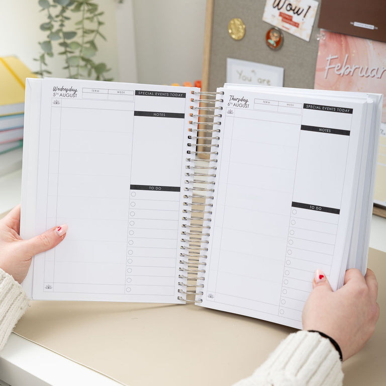Two open planners held by hands on a desk with a decorative wall in the background