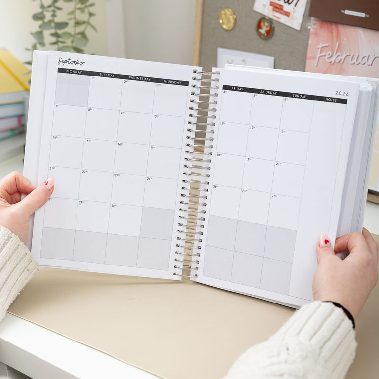 Person holding a spiral-bound planner with a calendar on a desk.