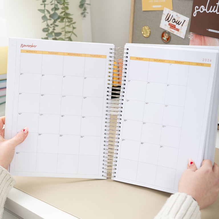 Person holding an open spiral-bound planner with a calendar on a desk.