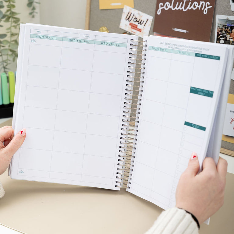 Person holding an open planner with a desk and motivational poster in the background