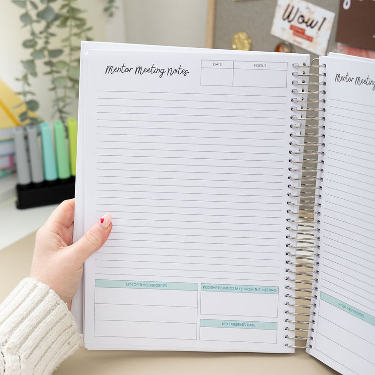 Person holding a spiral-bound planner with notes on a desk.