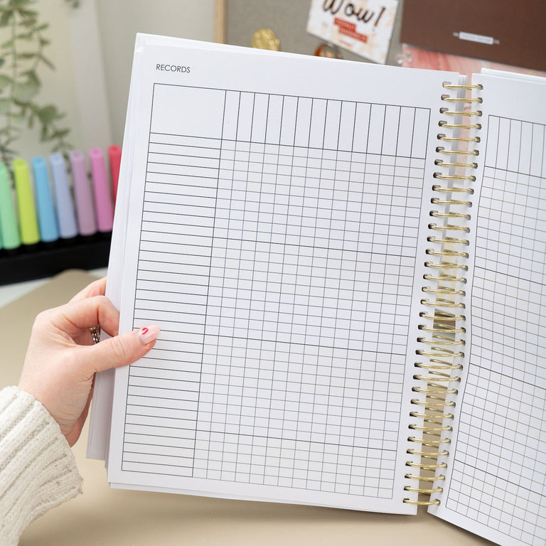 Person holding an open spiral-bound notebook with grid pages on a desk.