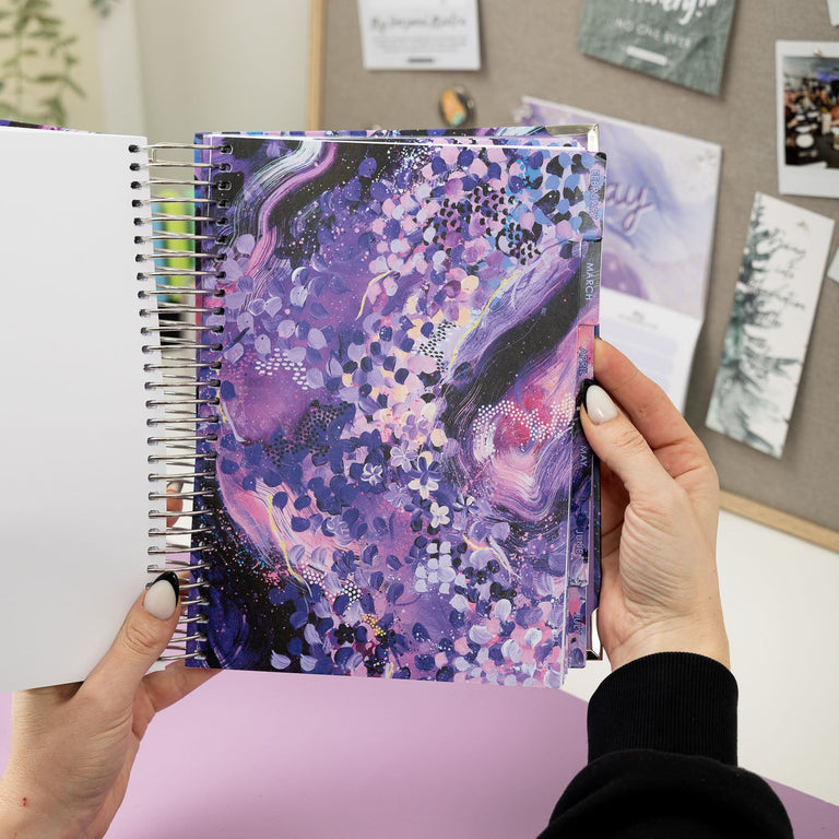 Person holding a spiral-bound notebook with a colorful cover against a wall with pinned photos.