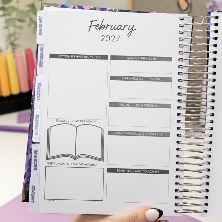 Person holding a planner with February 2027 layout on a desk with stationery items.