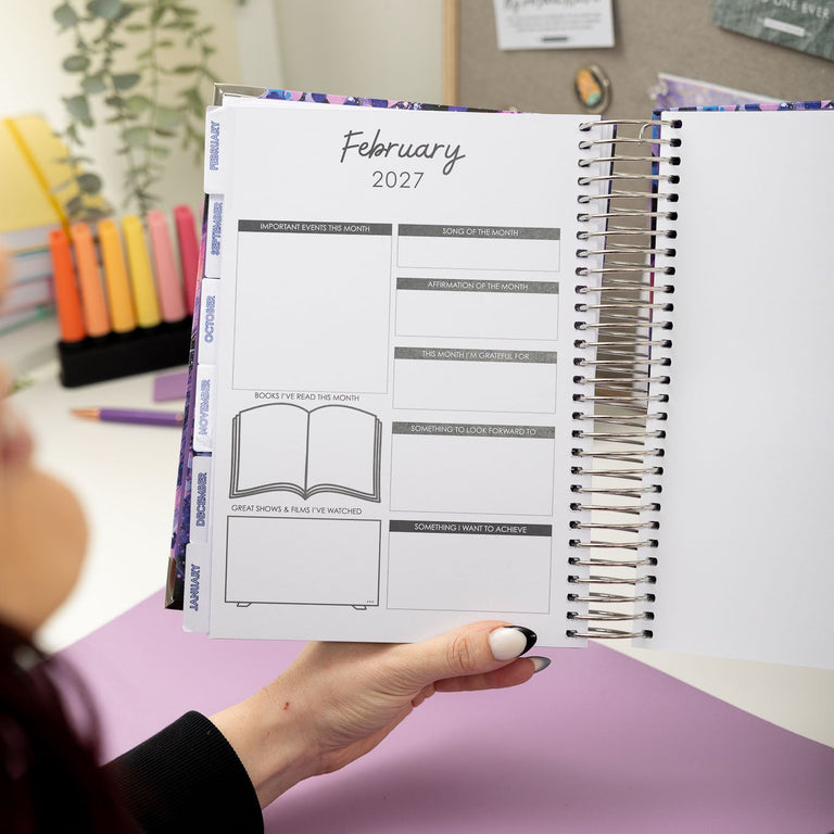 Person holding a planner with February 2027 layout on a desk with stationery items.