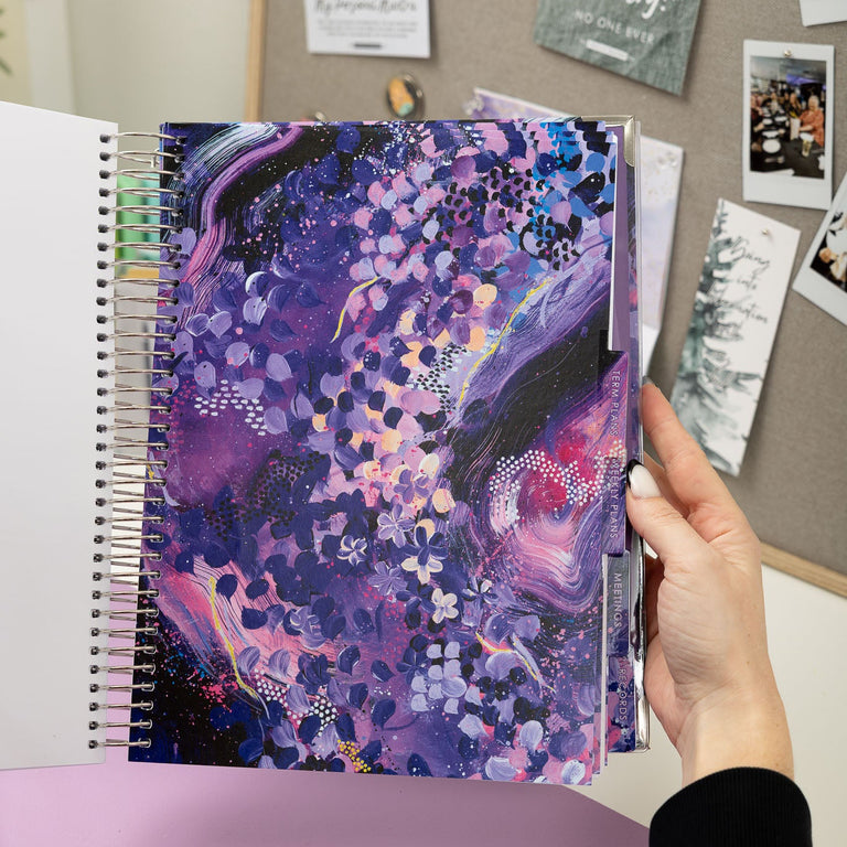 Person holding a spiral-bound notebook with a colorful purple cover against a wall with pinned photos and notes.