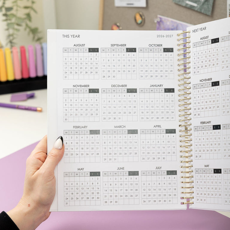 Person holding a spiral-bound planner with a calendar on a desk.