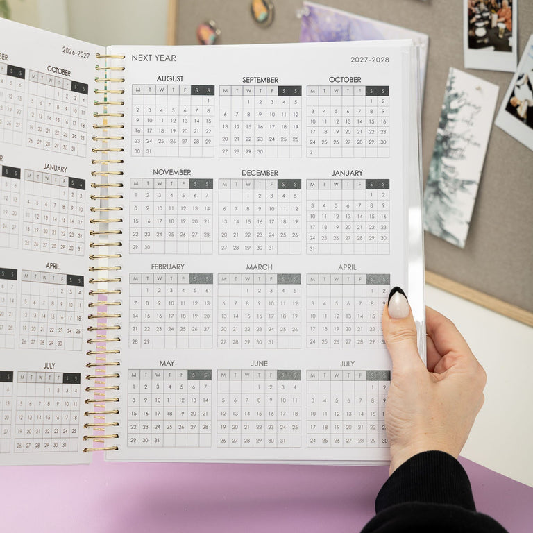 Person holding an open annual planner with a calendar view, set against a wall with photos and notes.