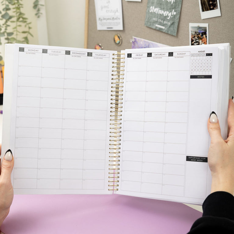 Person holding a spiral-bound planner with a pink surface and bulletin board in the background