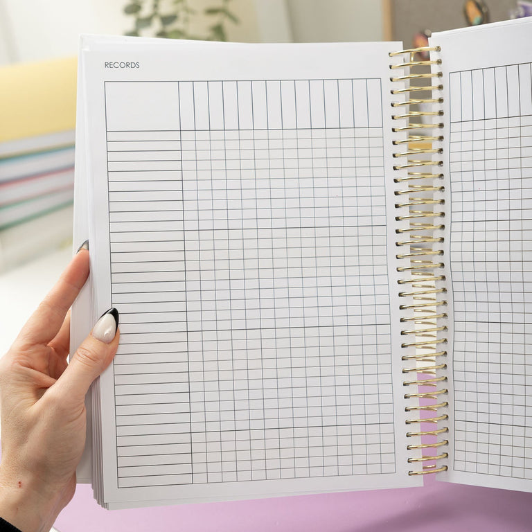 Person holding an open spiral-bound notebook with grid pages on a light purple surface.