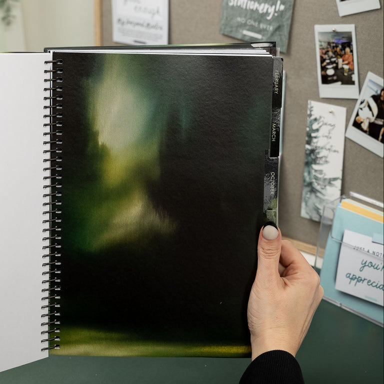 Person holding a spiral-bound notebook with a blurred green image on a desk with a bulletin board in the background.