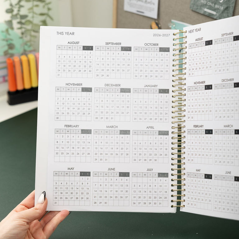 Person holding an open spiral-bound planner with a calendar on a green surface.