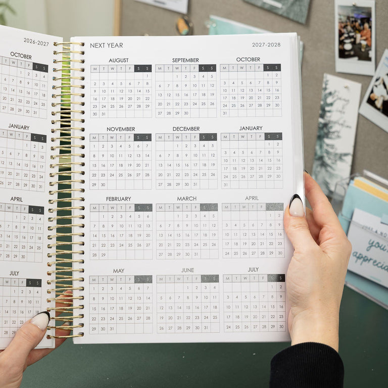 Person holding a spiral-bound planner with a calendar on a desk.
