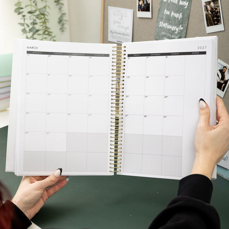 Person holding an open spiral-bound planner with a calendar on a desk.