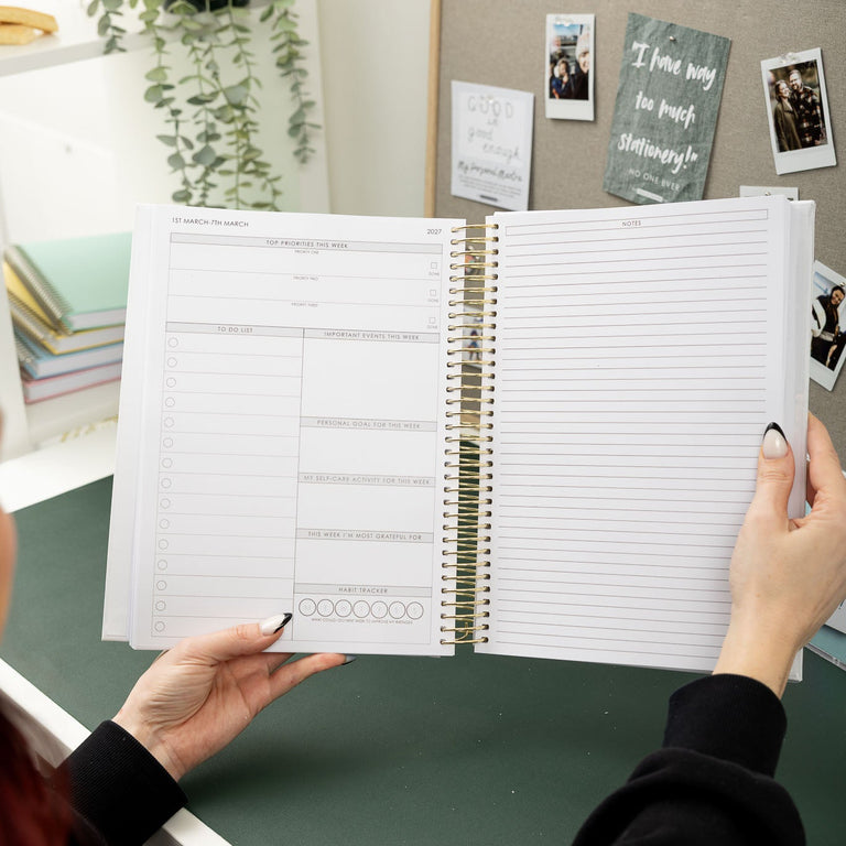 Person holding an open spiral-bound notebook with lined pages in a home office setting.