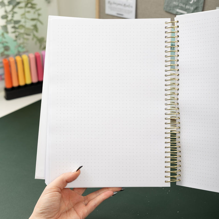 Open spiral-bound notebook held by a hand on a green surface with stationery items in the background.