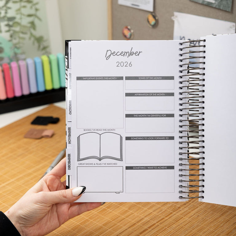 Person holding a planner open to December 2026 on a desk with a colorful calendar in the background.