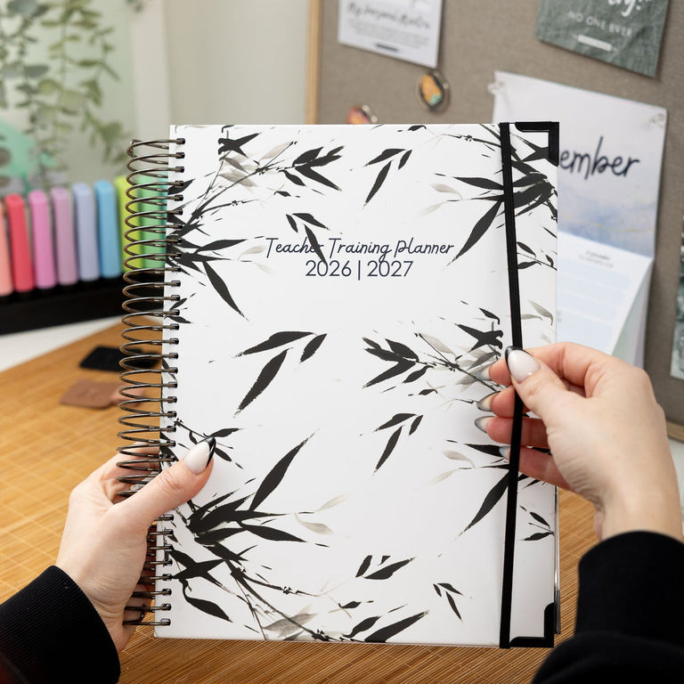 Person holding a Teacher Training Planner with a black and white leaf design on a desk.