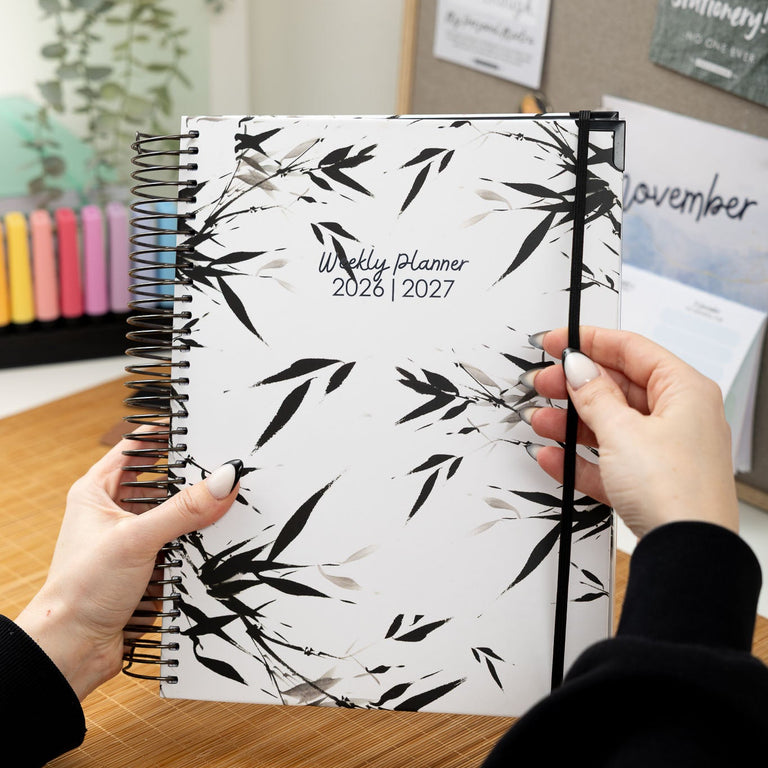 Person holding a weekly planner with a black and white leaf pattern, set against a blurred indoor background.