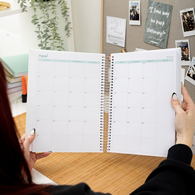 Person holding a spiral-bound planner open on a desk with a bulletin board in the background.