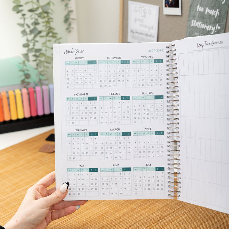 Person holding an open planner on a desk with a colorful calendar and plant in the background