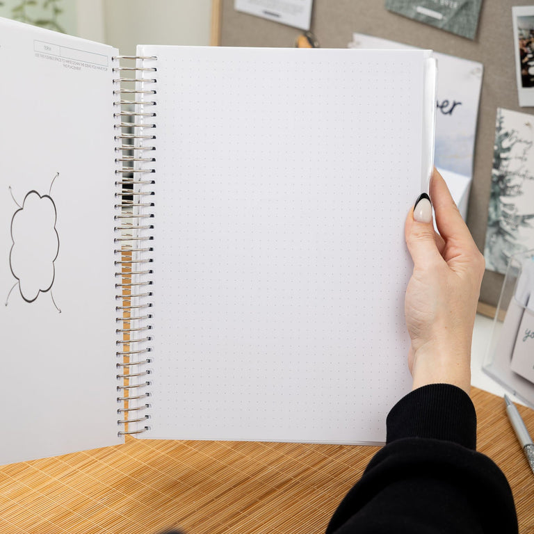 Person holding a spiral-bound notebook with a hand-drawn cloud on a desk.