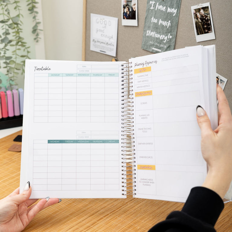 Person holding a spiral-bound planner open on a desk with a bulletin board in the background.