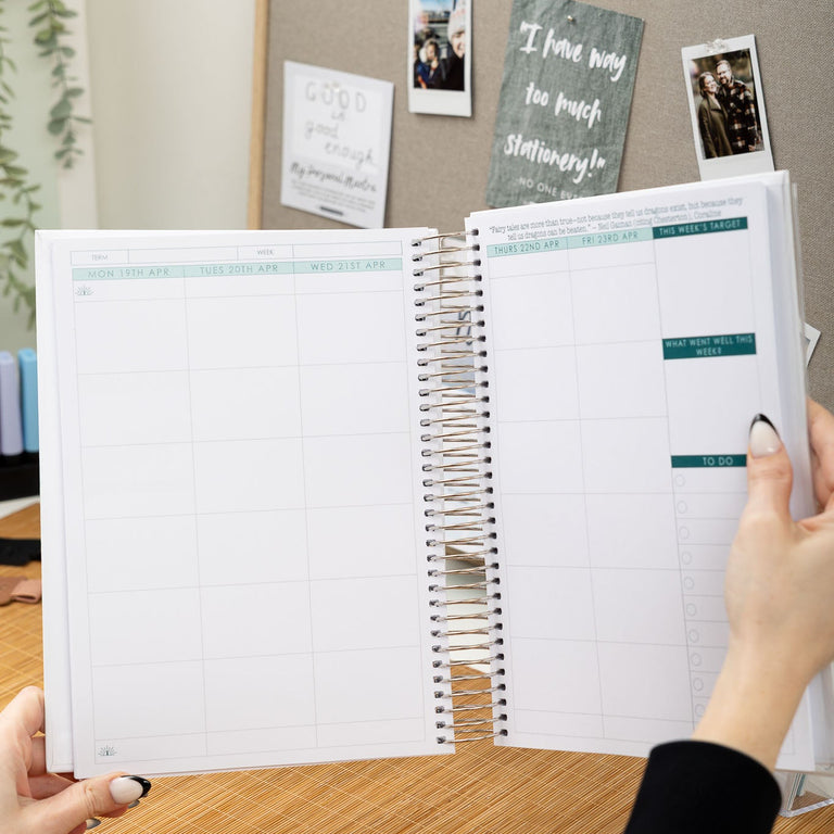 Person holding a planner open on a desk with a bulletin board in the background