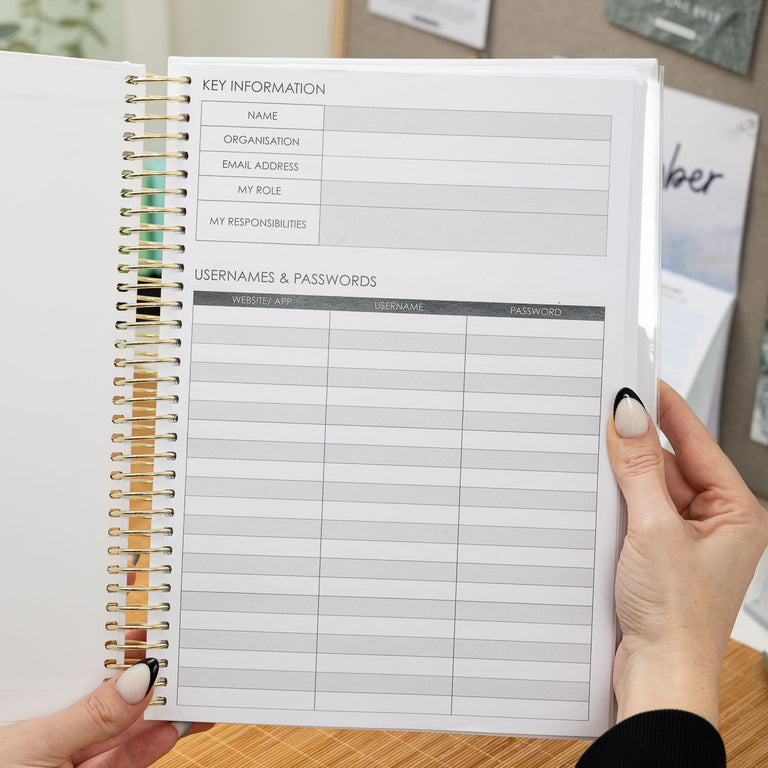 Person holding a spiral-bound notebook with a password list on a wooden surface.