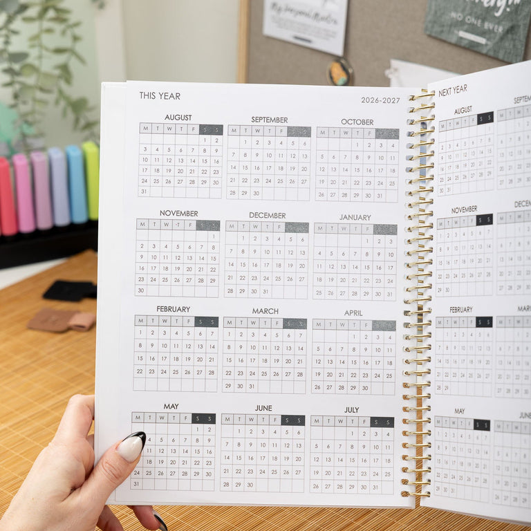 Person holding a spiral-bound planner with a calendar view, set against a desk and office background.