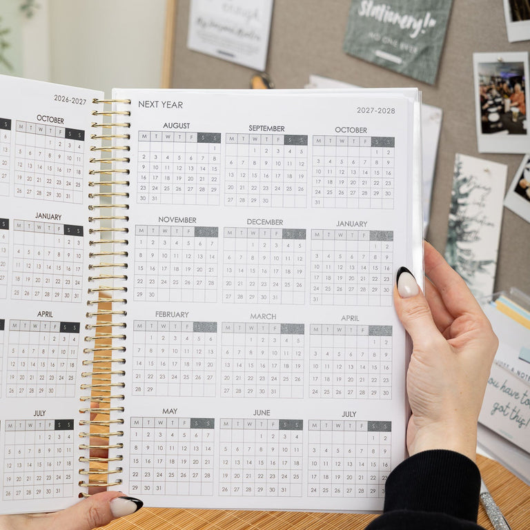 Person holding a spiral-bound planner with a calendar on a desk.