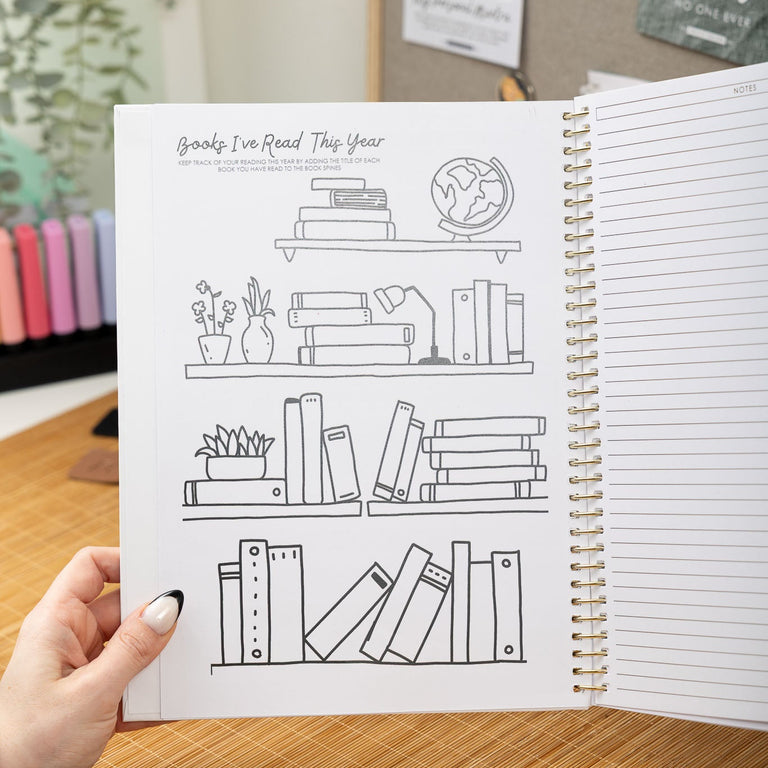 Open notebook with a page featuring line drawings of books on a shelf, held by a hand on a wooden surface.