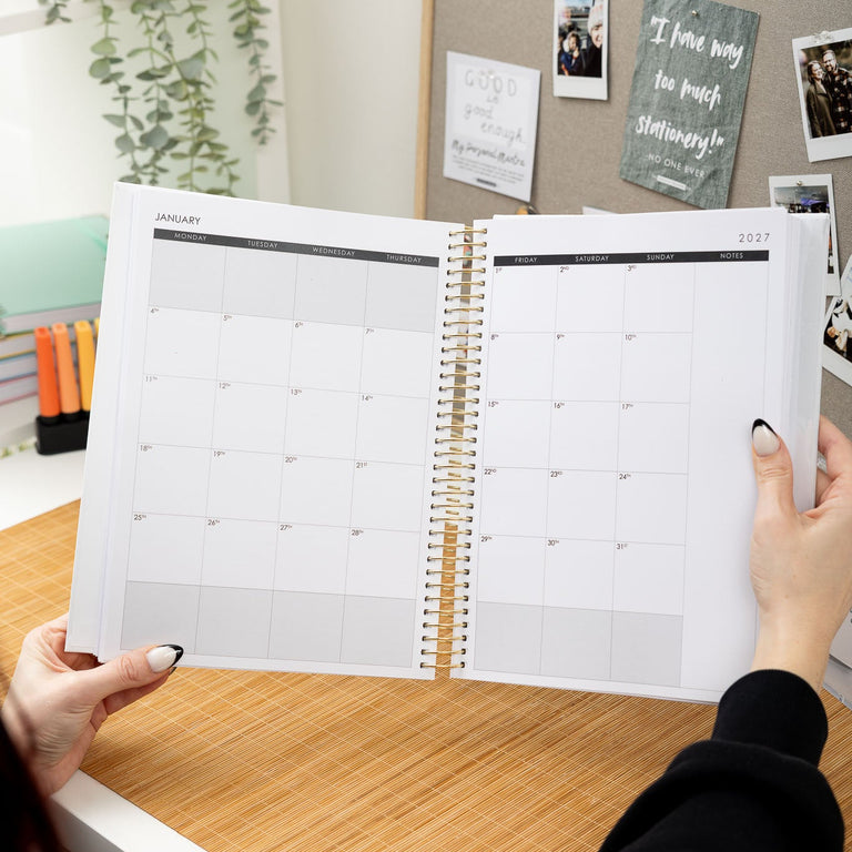 Person holding a spiral-bound planner open on a desk with a bulletin board in the background
