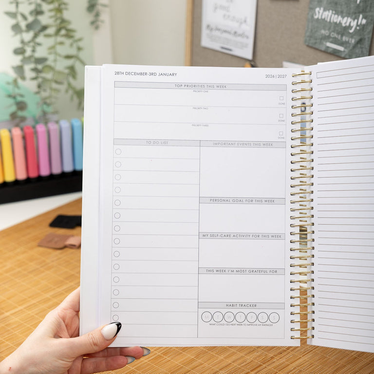Person holding a spiral-bound planner with a wooden desk and decorative items in the background