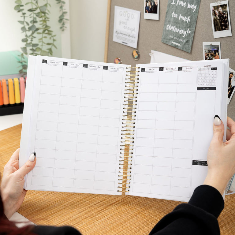 Person holding an open spiral-bound planner on a desk with a bulletin board in the background.