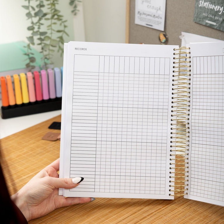 Person holding a spiral-bound notebook with grid paper on a desk with stationery items.