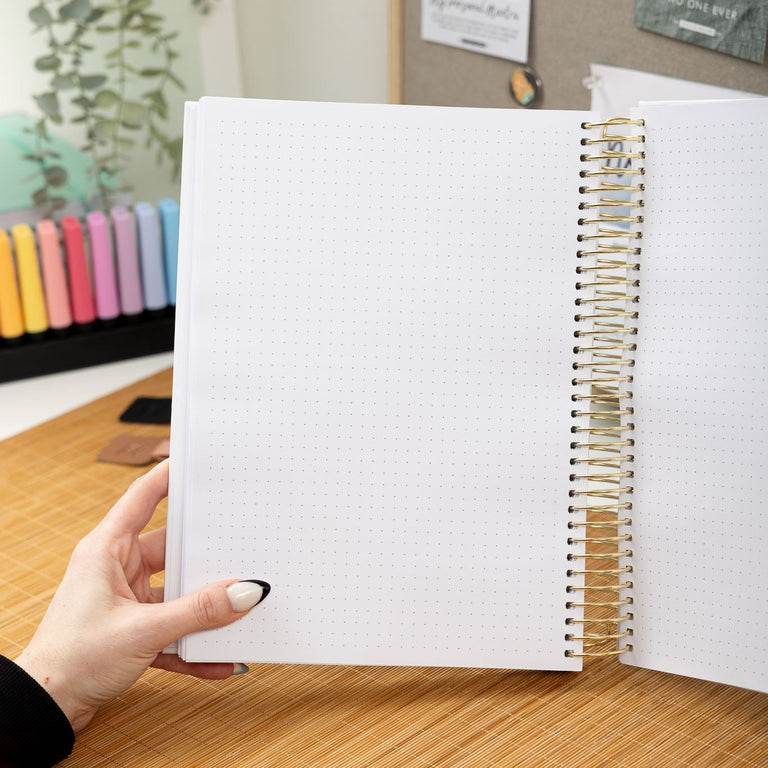Open notebook held by a person on a wooden desk with a colorful pencil holder in the background.