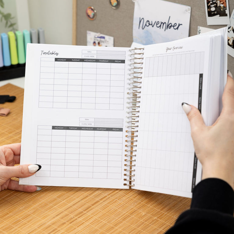 Person holding a spiral-bound planner open on a wooden desk with a blurred background