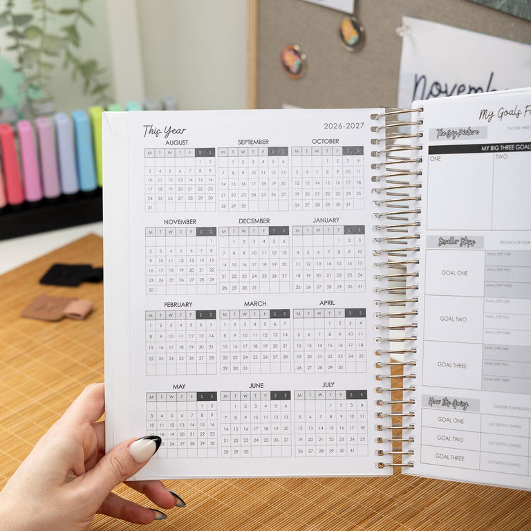 Person holding a spiral-bound planner with a calendar on a wooden desk.