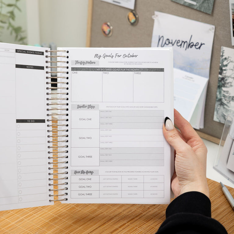 Person holding a spiral-bound planner with a wooden desk and bulletin board in the background