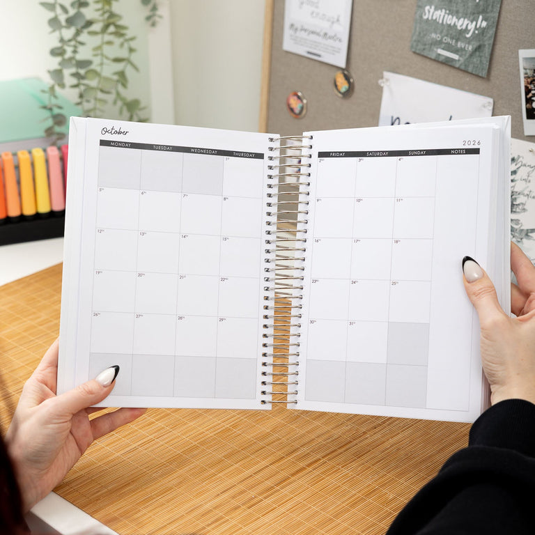 Person holding a spiral-bound planner open on a desk with a bulletin board in the background