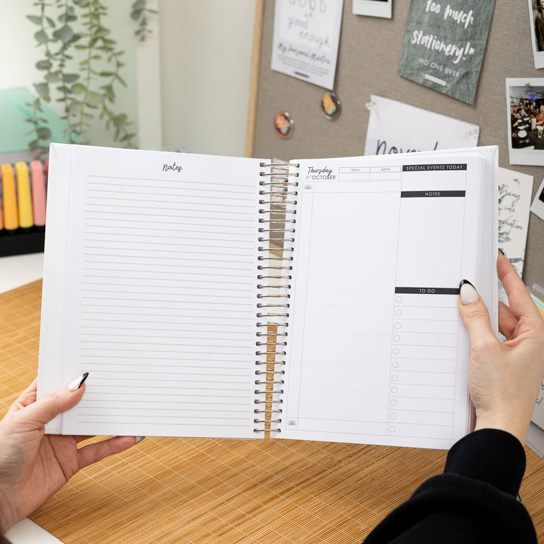 Person holding a spiral-bound planner with a desk and bulletin board in the background