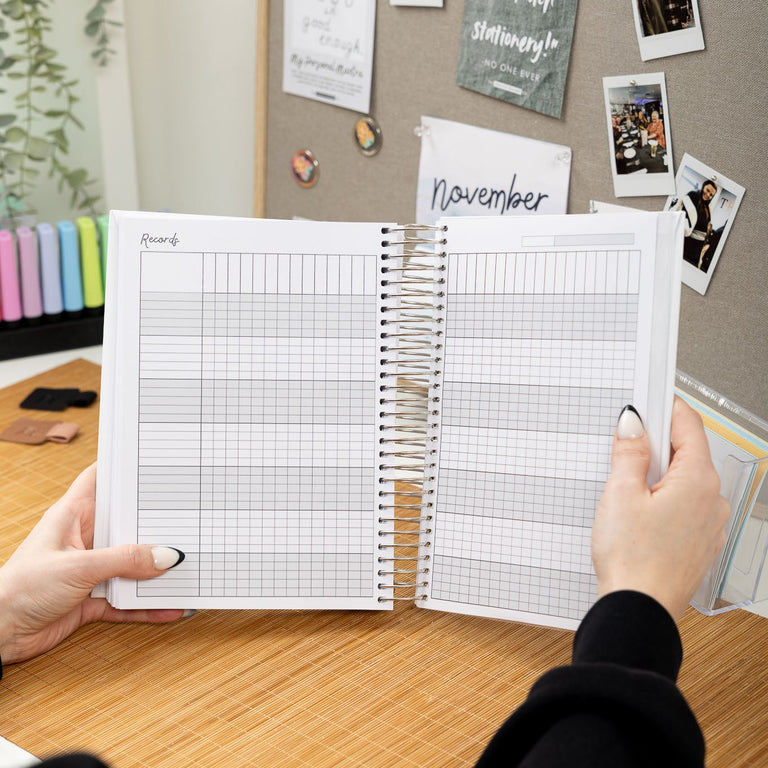 Person holding a planner with a grid layout on a desk with photos and notes on the wall.