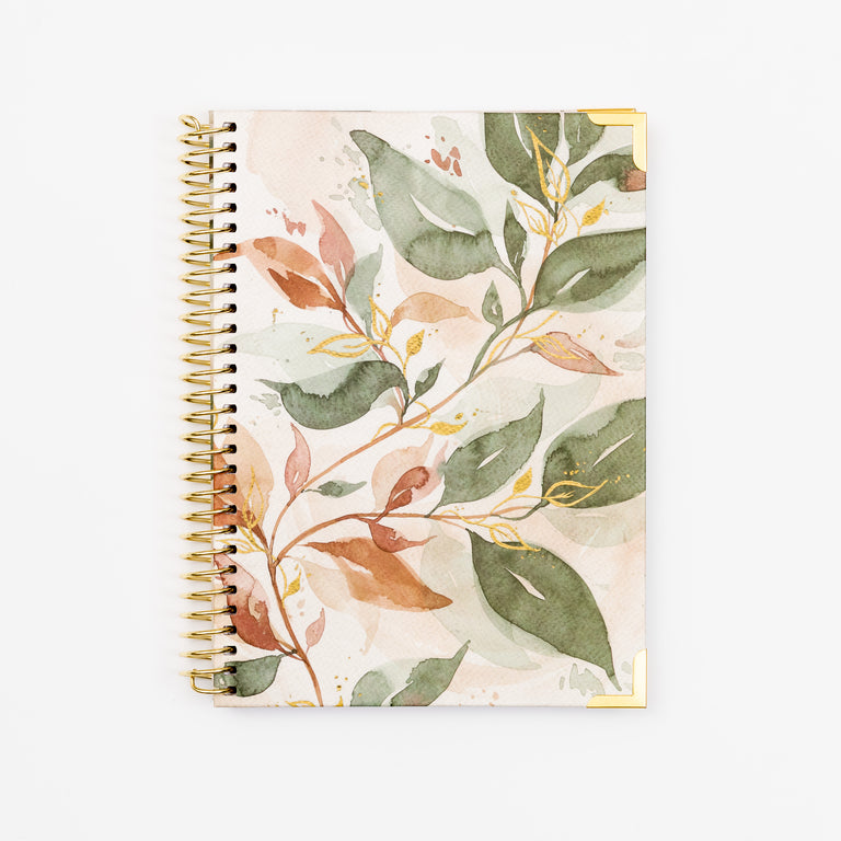 A spiral-bound notebook with a watercolor botanical cover featuring green leaves and warm brown and peach branches on a light cream background, bound with gold-toned metal rings.