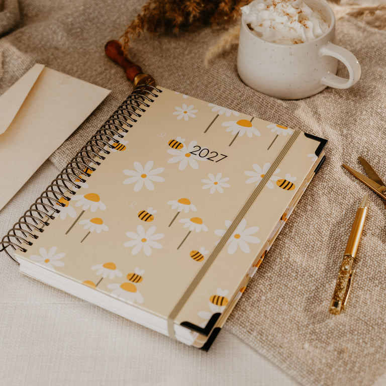 A spiral-bound 2027 planner with a beige cover decorated with white daisies and yellow bees, styled on a beige blanket next to a white mug of hot chocolate, gold scissors, and dried pampas grass.