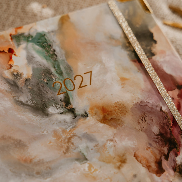 A close-up of a 2027 spiral-bound planner with an abstract marbled cover in cream, beige, brown, and muted green tones, featuring a gold glittery edge and gold '2027' text.