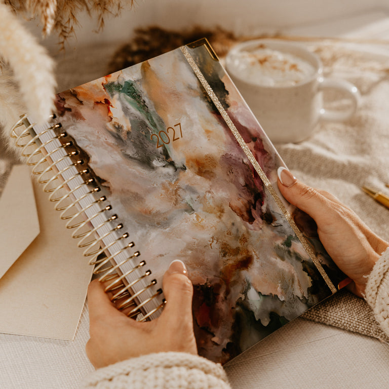 A person holds a 2027 spiral-bound planner with an abstract watercolor cover in a cozy, warm-toned setting with a mug and dried grass.