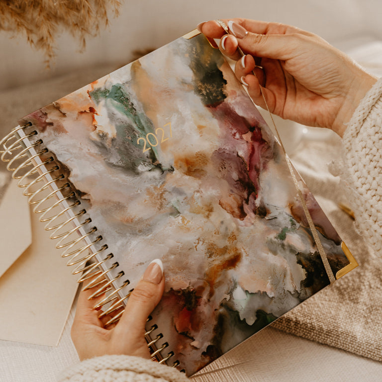 A person holds a spiral-bound planner with an abstract watercolor cover in warm beige, blush, green, and brown tones. The scene is styled with a cream knit sweater, a beige pillow, and dried pampas grass in the background.